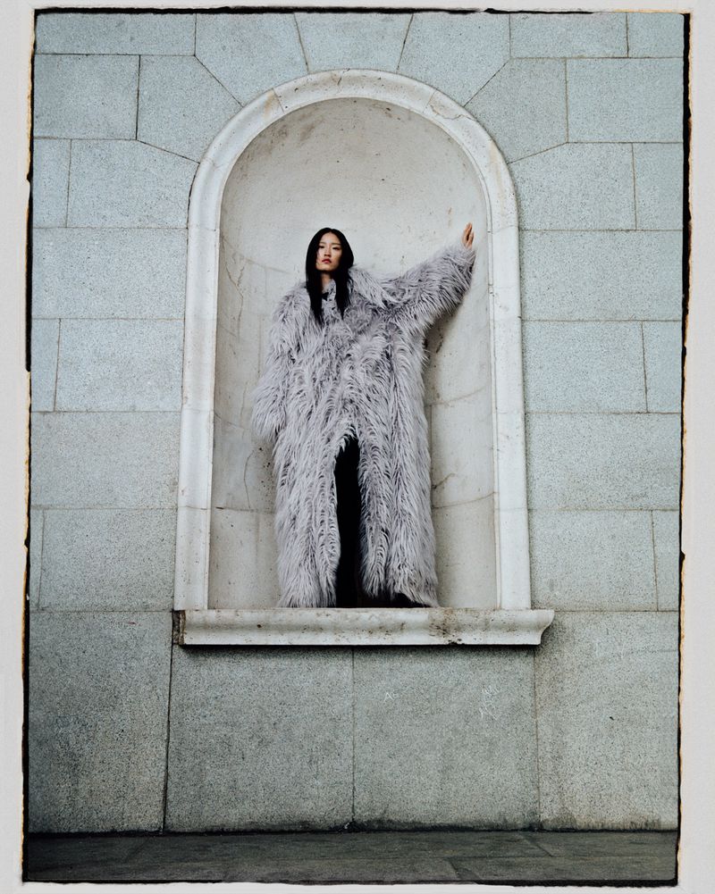 Elegant woman poses in a stone alcove wearing a long grey faux fur coat, creating a striking fashion editorial image.