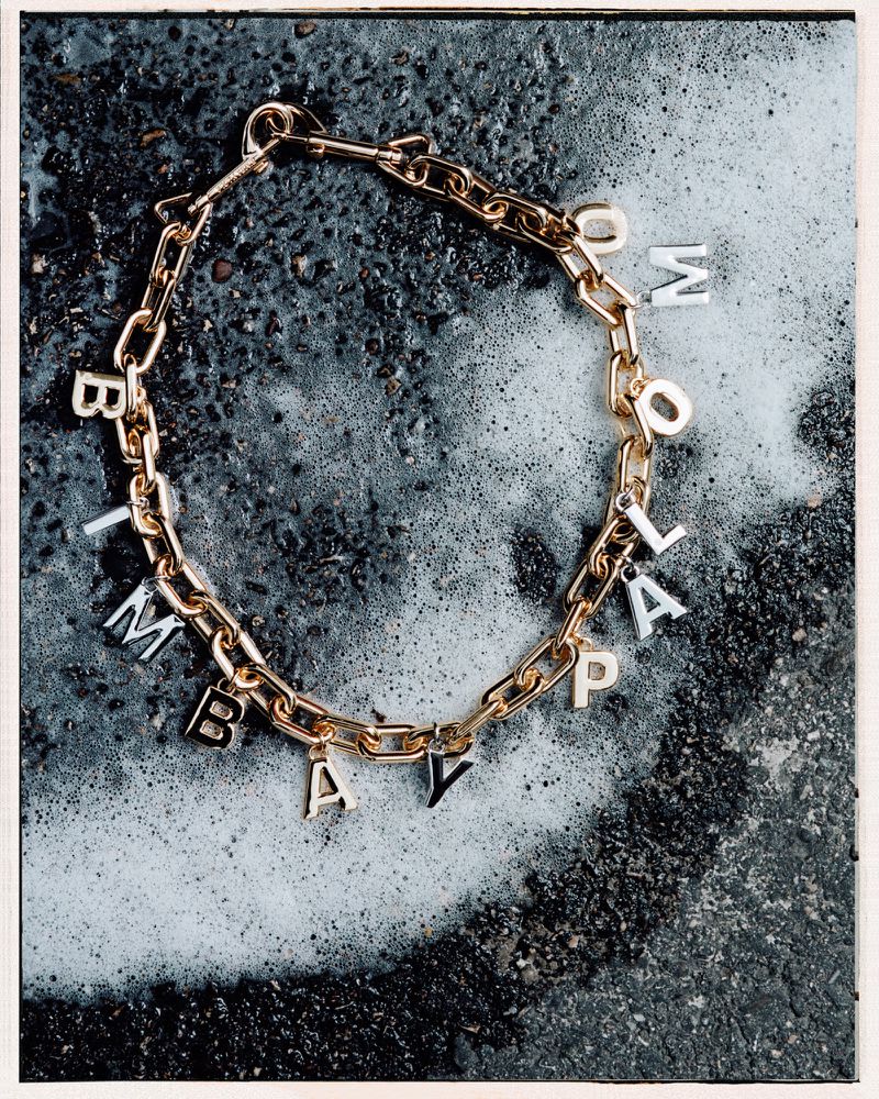 Gold chain link necklace with letter charms spelling "TOMMOLAPAY" on a dark textured background with white accents.