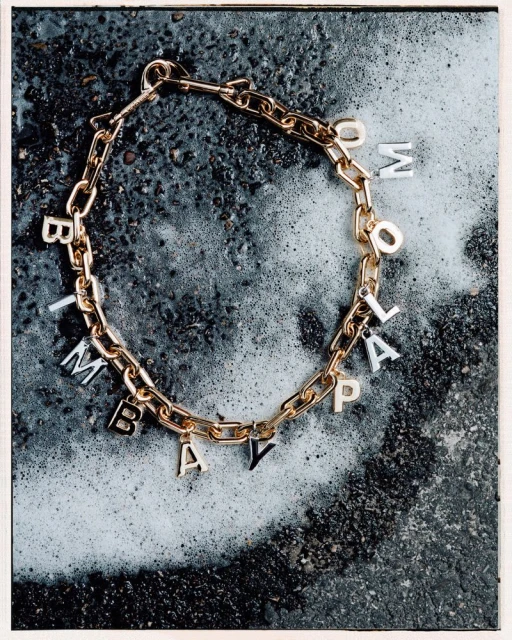 Gold chain link necklace with letter charms spelling "TOMMOLAPAY" on a dark textured background with white accents.