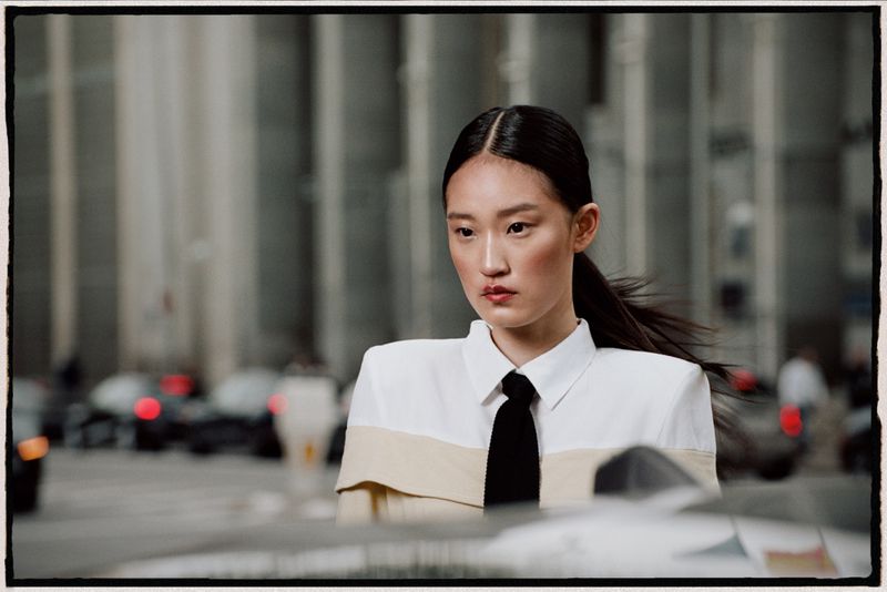 Fashion portrait of a young Asian woman wearing a white shirt and black tie, posing in a city street with blurred background.