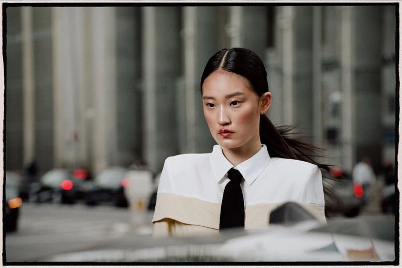 Fashion portrait of a young Asian woman wearing a white shirt and black tie, posing in a city street with blurred background.