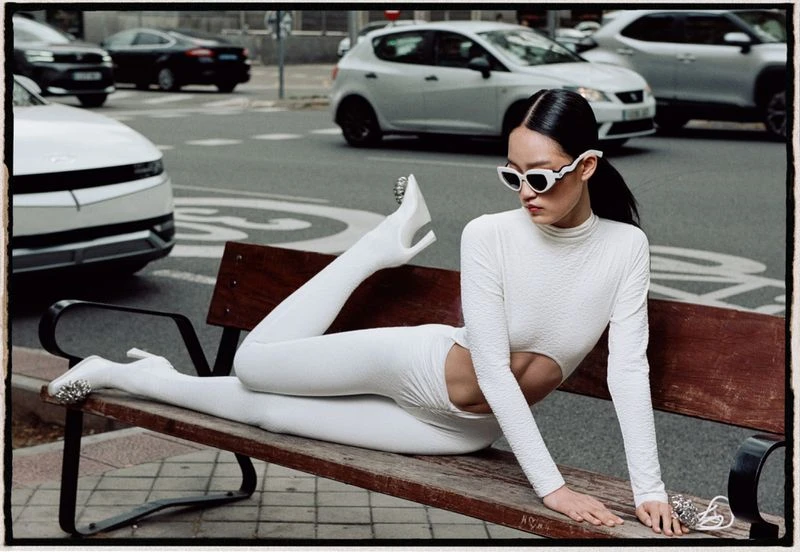Fashion shot of a woman in a white bodysuit and sunglasses reclining on a bench in a city street with cars in the background.