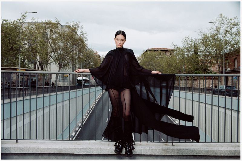 Fashion shot of a young Asian woman wearing a long black gown with cape, posing on a bridge with city buildings and trees in background.