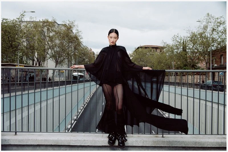 Fashion shot of a young Asian woman wearing a long black gown with cape, posing on a bridge with city buildings and trees in background.