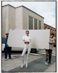 Model in white long-sleeve crop top and leggings standing outdoors while two crew members hold a large white board behind her