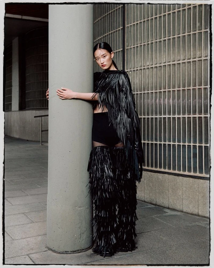 Model wearing a black fringed outfit hugging a concrete pillar outdoors for Ladygunn Magazine
