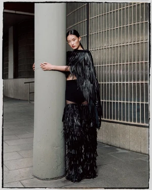 Model wearing a black fringed outfit hugging a concrete pillar outdoors for Ladygunn Magazine