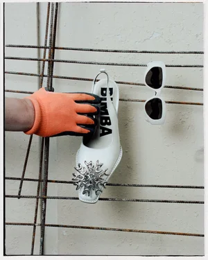 Hand in orange glove holding a white slingback shoe with silver jewel embellishment near white sunglasses on rebar grid.