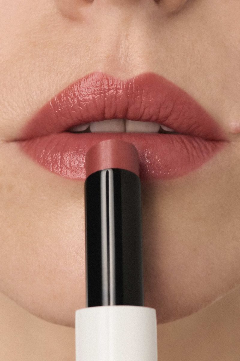 Close-up of a model applying rose-tinted lipstick from a white and black tube onto lips