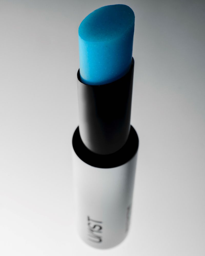 Close-up product shot of a blue lip balm tube with a sleek silver and black container against a minimalist white gradient background.