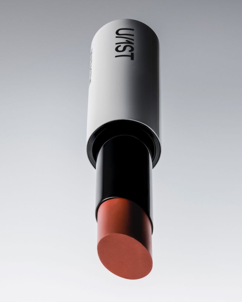 Close-up product shot of an open U/1ST lip balm tube with a peach-toned stick against a grey seamless background