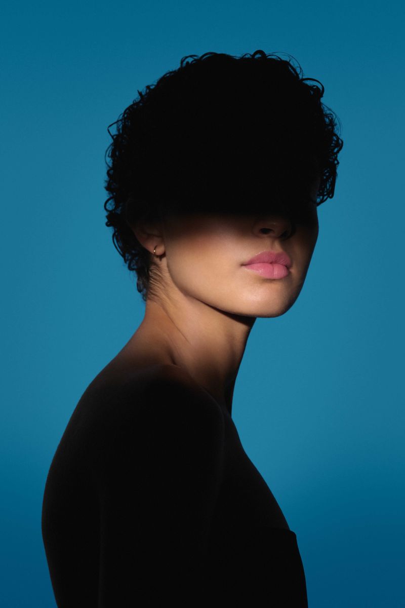 Studio portrait of a young woman with an afro hairstyle against a light blue backdrop, face partially obscured by shadow.