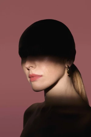 Close-up portrait of a woman wearing a black cap casting a shadow over her eyes against a muted pink background.
