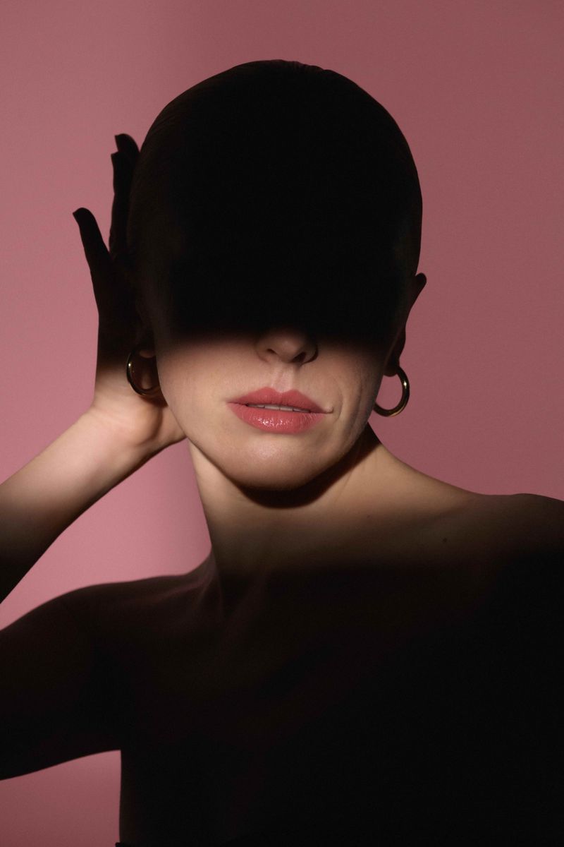 Close-up portrait of a woman with a heavily shadowed face against a pink background, wearing hoop earrings and pink lipstick.