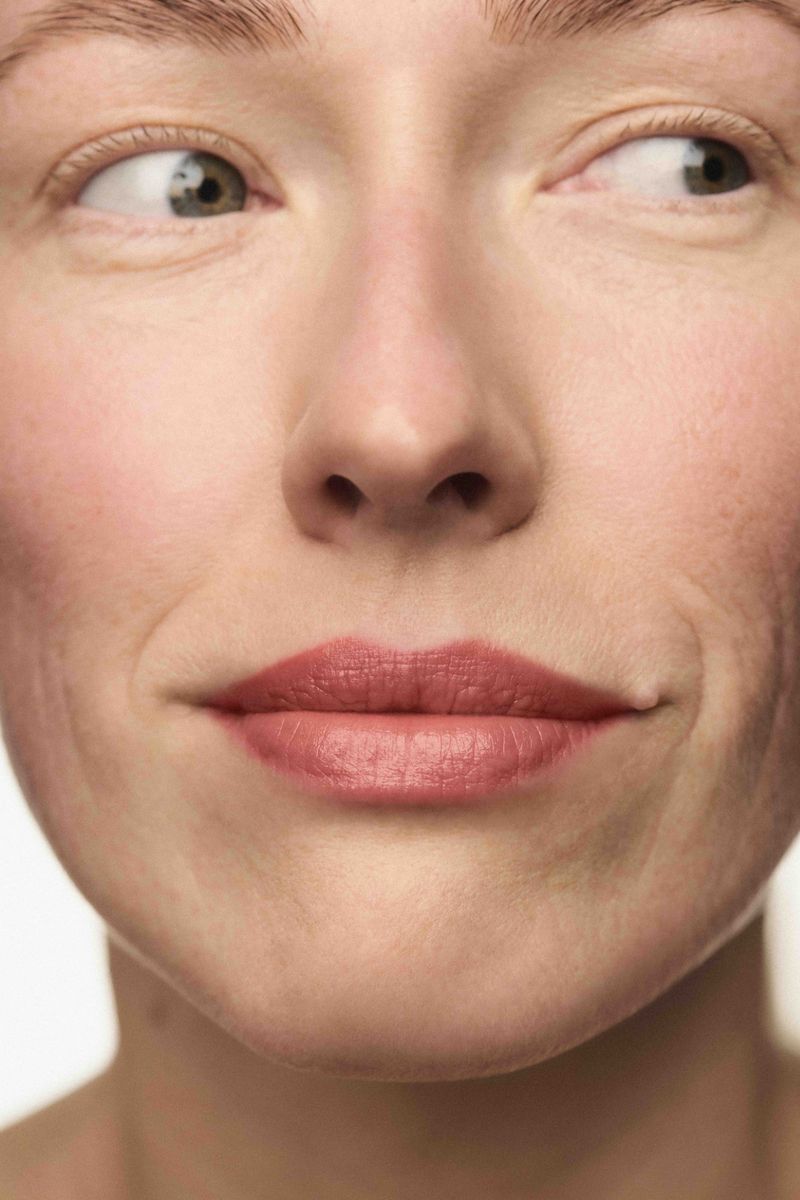 Close-up portrait of a woman wearing dusty rose lipstick looking sideways against a neutral background.