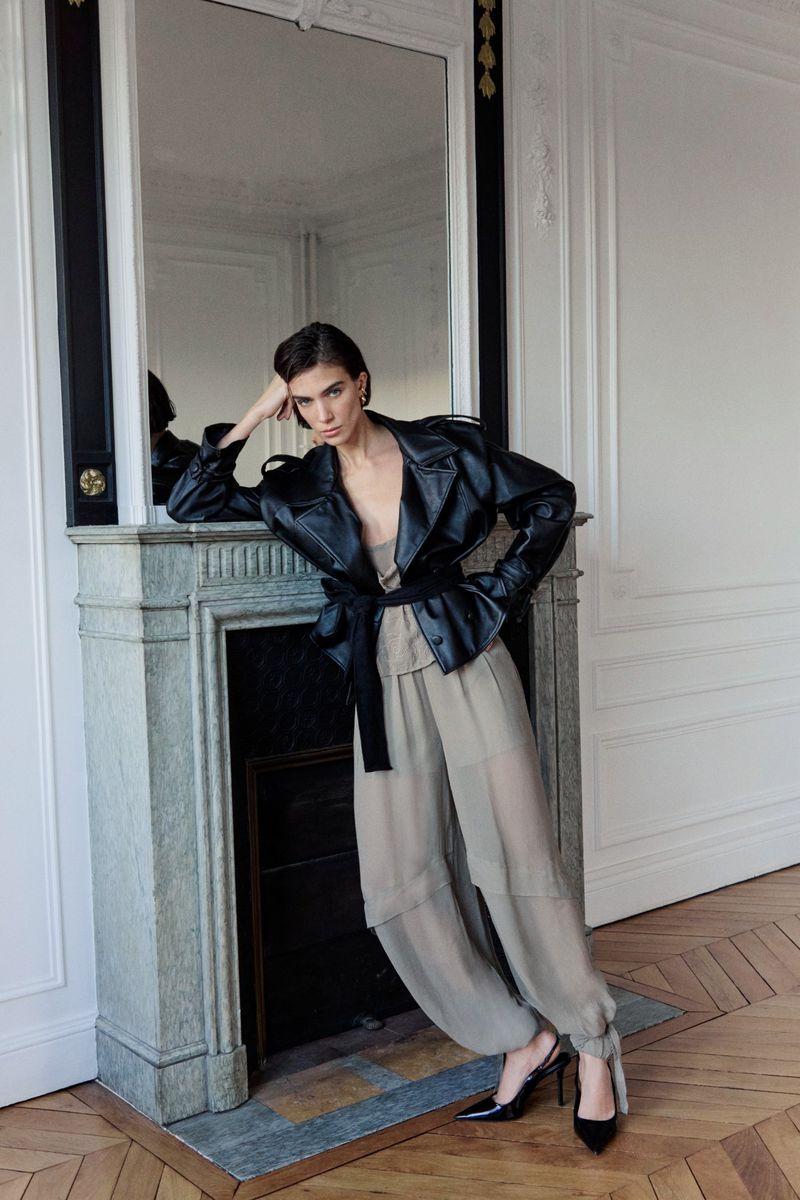Model leaning against a marble fireplace wearing a black leather jacket and beige trousers in an interior room.