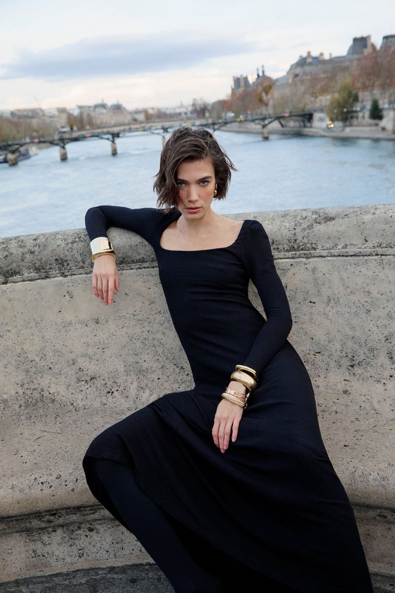Woman in long black dress wearing gold cuffs leaning against a stone bridge over a river in Paris