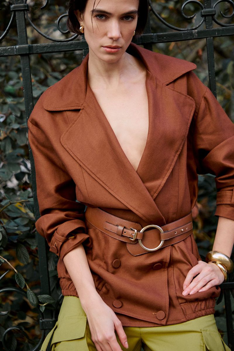 Model in brown belted jacket and olive green trousers leaning against an ornate dark iron fence