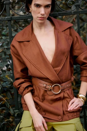 Model in brown belted jacket and olive green trousers leaning against an ornate dark iron fence