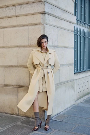 Model wearing a long beige belted trench coat leaning against a stone building exterior wall on a paved street.