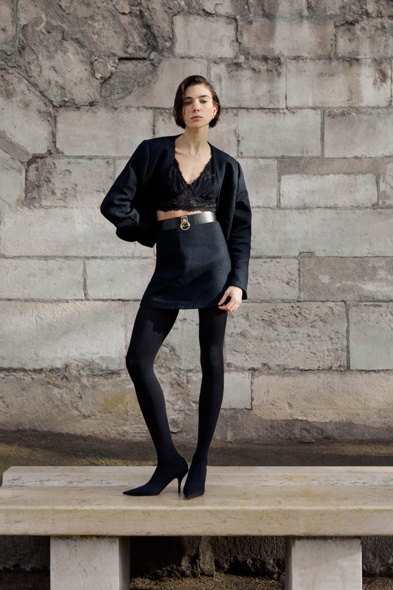 Model standing on a stone bench in front of a weathered brick wall wearing a dark jacket, black lace crop top, and skirt.