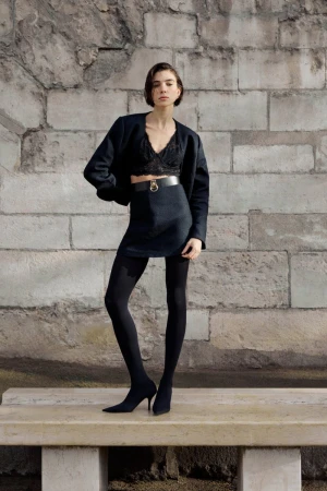 Model standing on a stone bench in front of a weathered brick wall wearing a dark jacket, black lace crop top, and skirt.
