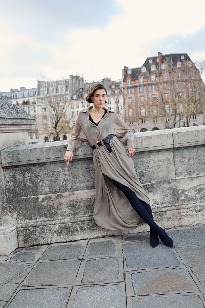 Woman in a long grey belted dress leaning against a stone bridge with historic buildings in the background