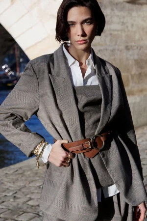 Model with dark hair wearing a grey plaid blazer and brown leather belt against an outdoor stone background