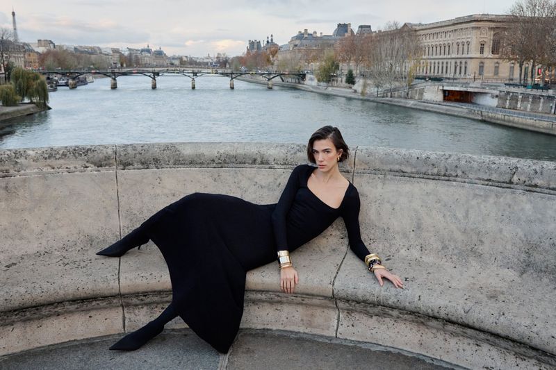 Woman in black long-sleeve dress reclining on a stone bridge overlooking a river and classical buildings.
