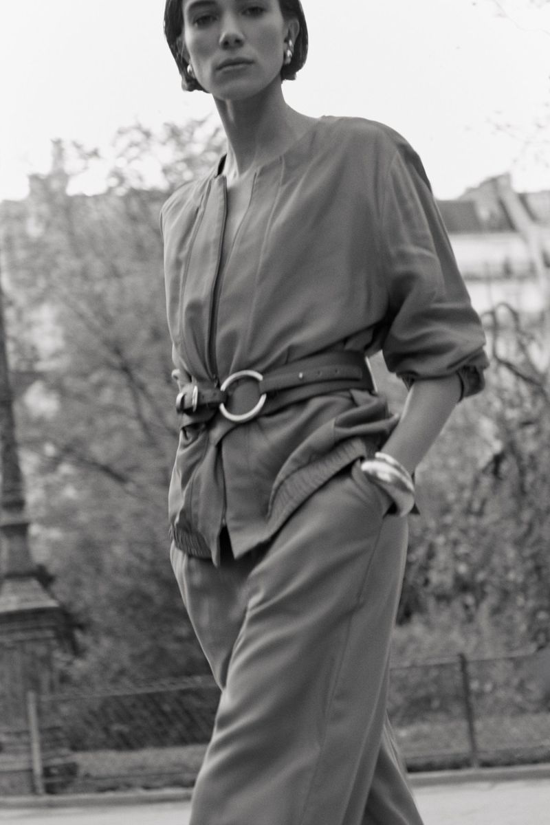 Model in belted jacket and trousers walking outdoors in a black and white fashion editorial photograph by Thomas Tebet.