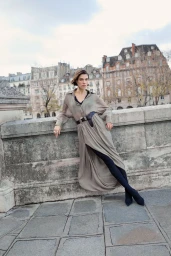 Woman in a long grey belted dress leaning against a stone bridge with historic buildings in the background