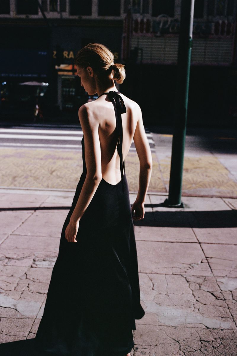 Model with hair in a low bun wearing a black halter dress walking on a city street.