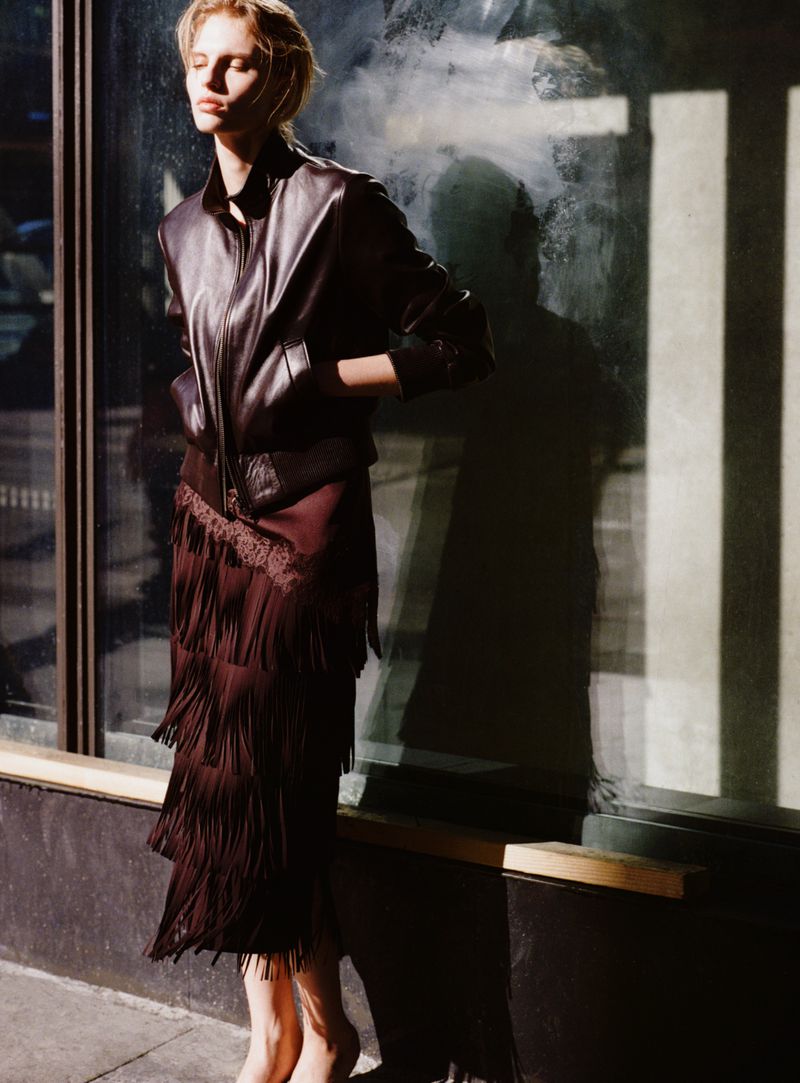 Model wearing a dark leather jacket and fringed skirt standing outdoors against a storefront window.