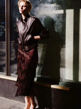 Model wearing a dark leather jacket and fringed skirt standing outdoors against a storefront window.
