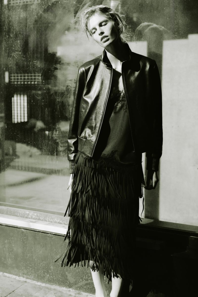 Model in a black leather jacket and fringed skirt posing outdoors against a textured window backdrop