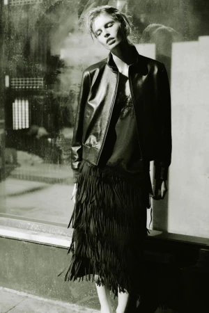 Model in a black leather jacket and fringed skirt posing outdoors against a textured window backdrop