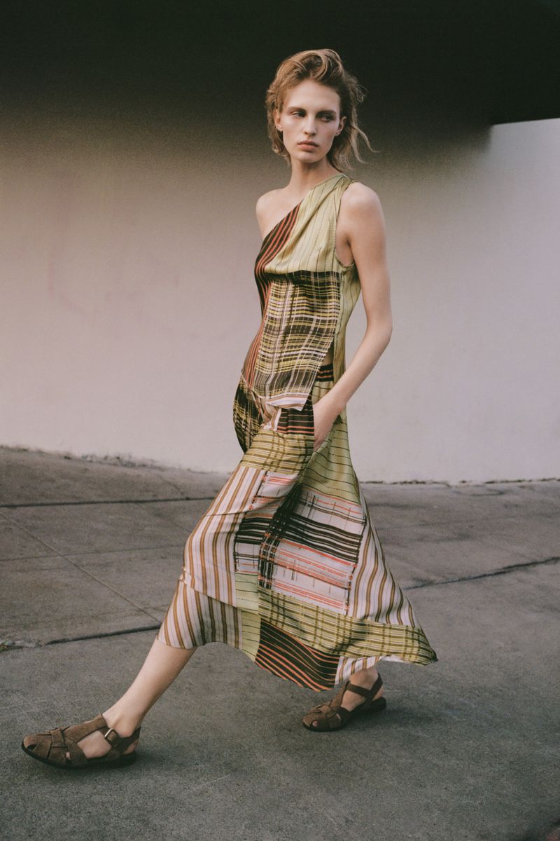 Model in a patterned one-shoulder dress walking outdoors against a neutral concrete backdrop