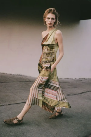 Model in a patterned one-shoulder dress walking outdoors against a neutral concrete backdrop