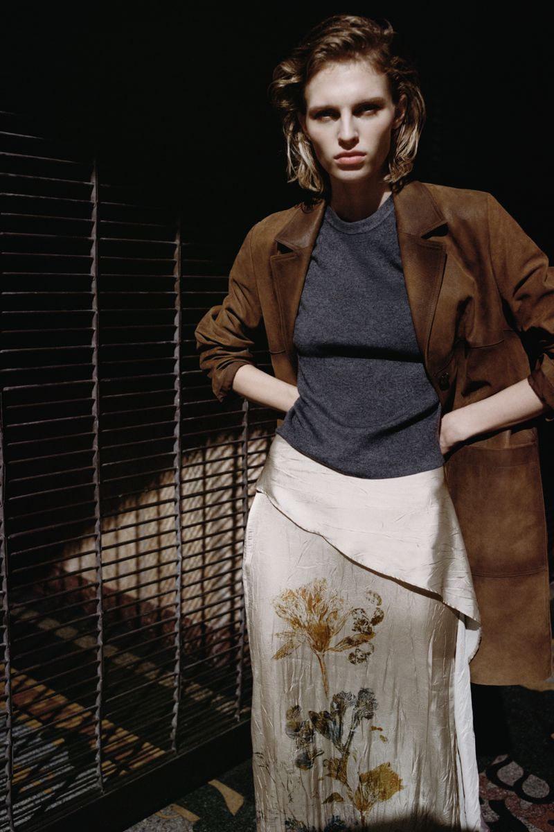 Model in brown suede coat, grey top and patterned silk skirt posing indoors by a metal structure