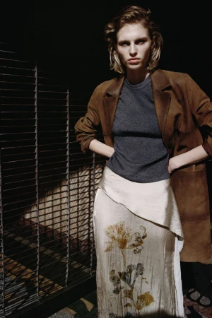Model in brown suede coat, grey top and patterned silk skirt posing indoors by a metal structure