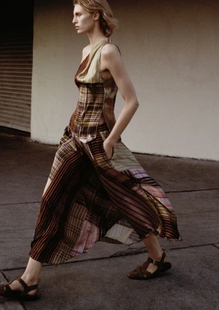 A fair-skinned woman walks outdoors in a flowy patterned dress and sandals on a concrete surface near a cream-colored building.