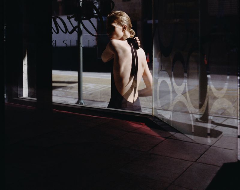 Model in a black backless halter dress viewed through a storefront window with graphic circular patterns.