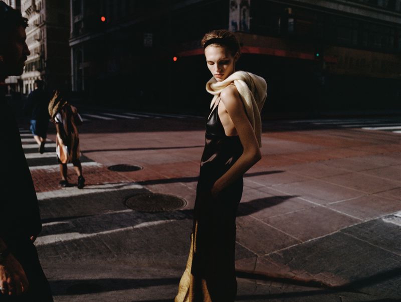 Model standing on a city street at dusk wearing a dark dress with a light sweater draped over shoulders