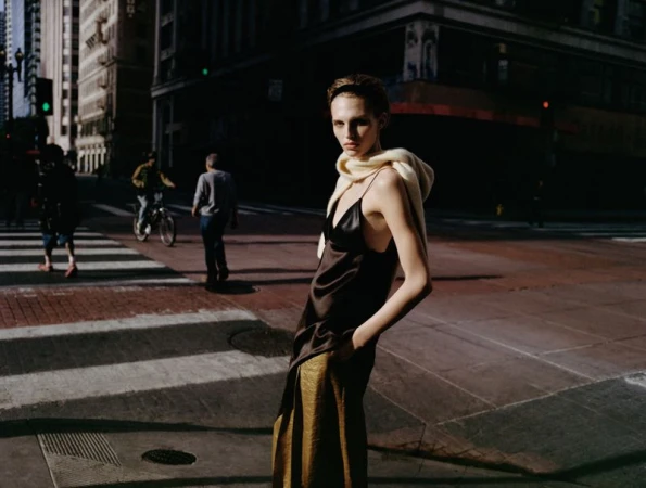 Model wearing a dark slip dress and draped cream sweater standing on a city street intersection at dusk.