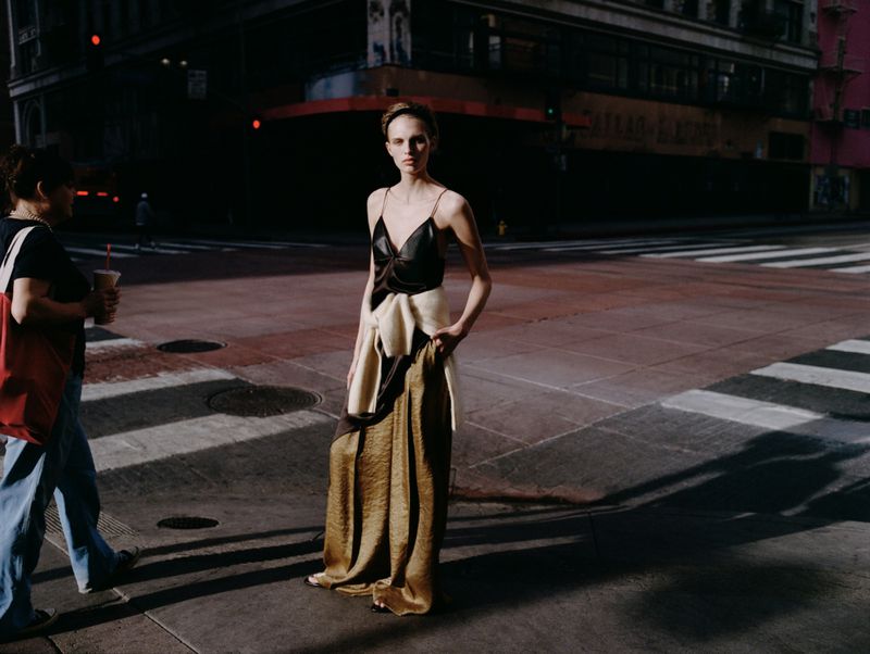 Model standing on a city street wearing a black top and long metallic skirt with a sweater tied around her waist.