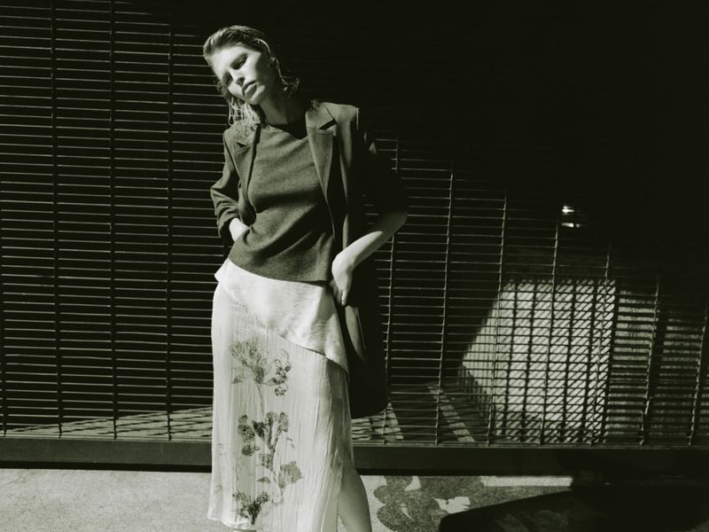 Model wearing a dark blazer, top, and floral skirt posing against a slatted metal gate for Zara.