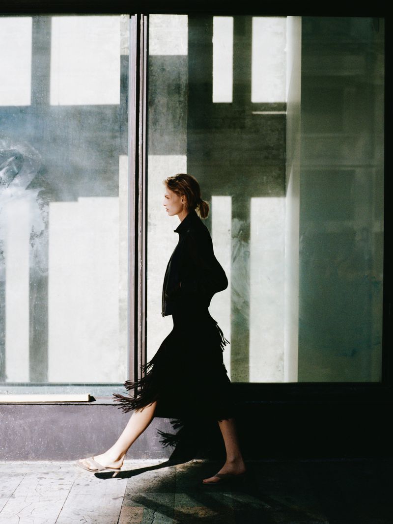 Model in profile walking past large glass windows wearing a black fringed skirt and dark jacket for Zara