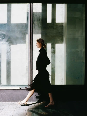 Model in profile walking past large glass windows wearing a black fringed skirt and dark jacket for Zara