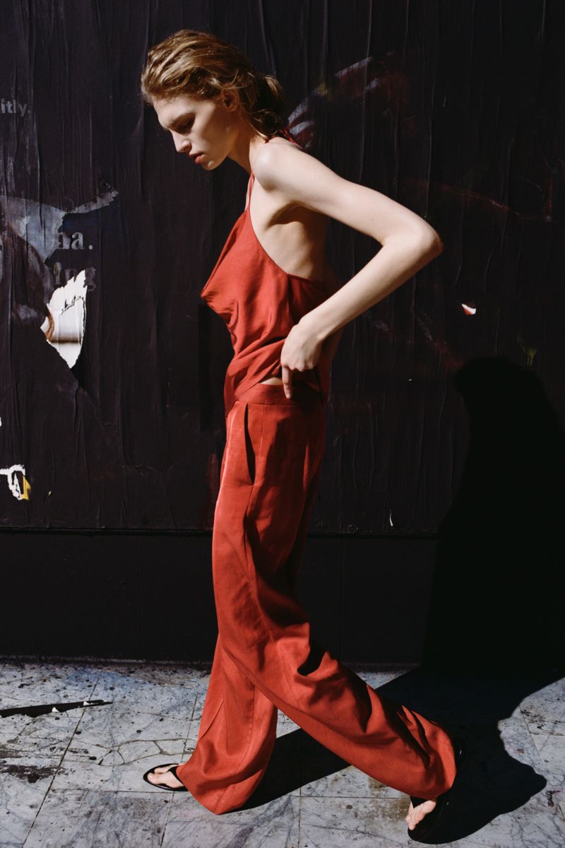 Model in a red silk top and matching trousers walking against a dark poster-covered wall for Zara