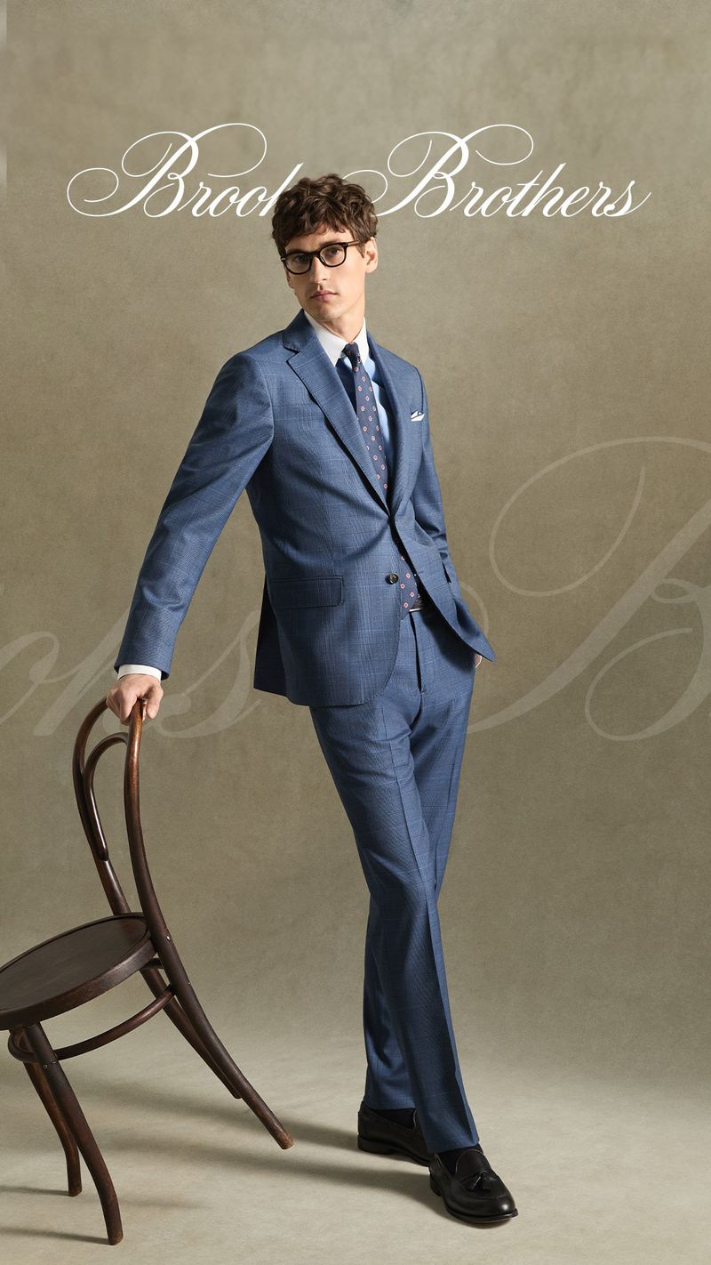 Male model in a blue suit and glasses leaning against a dark wooden chair against a neutral textured background
